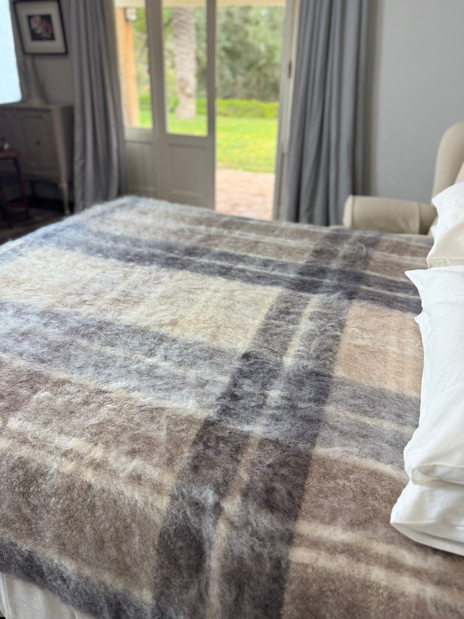 Plaid blanket on a bed in a room with a window showing greenery outside.