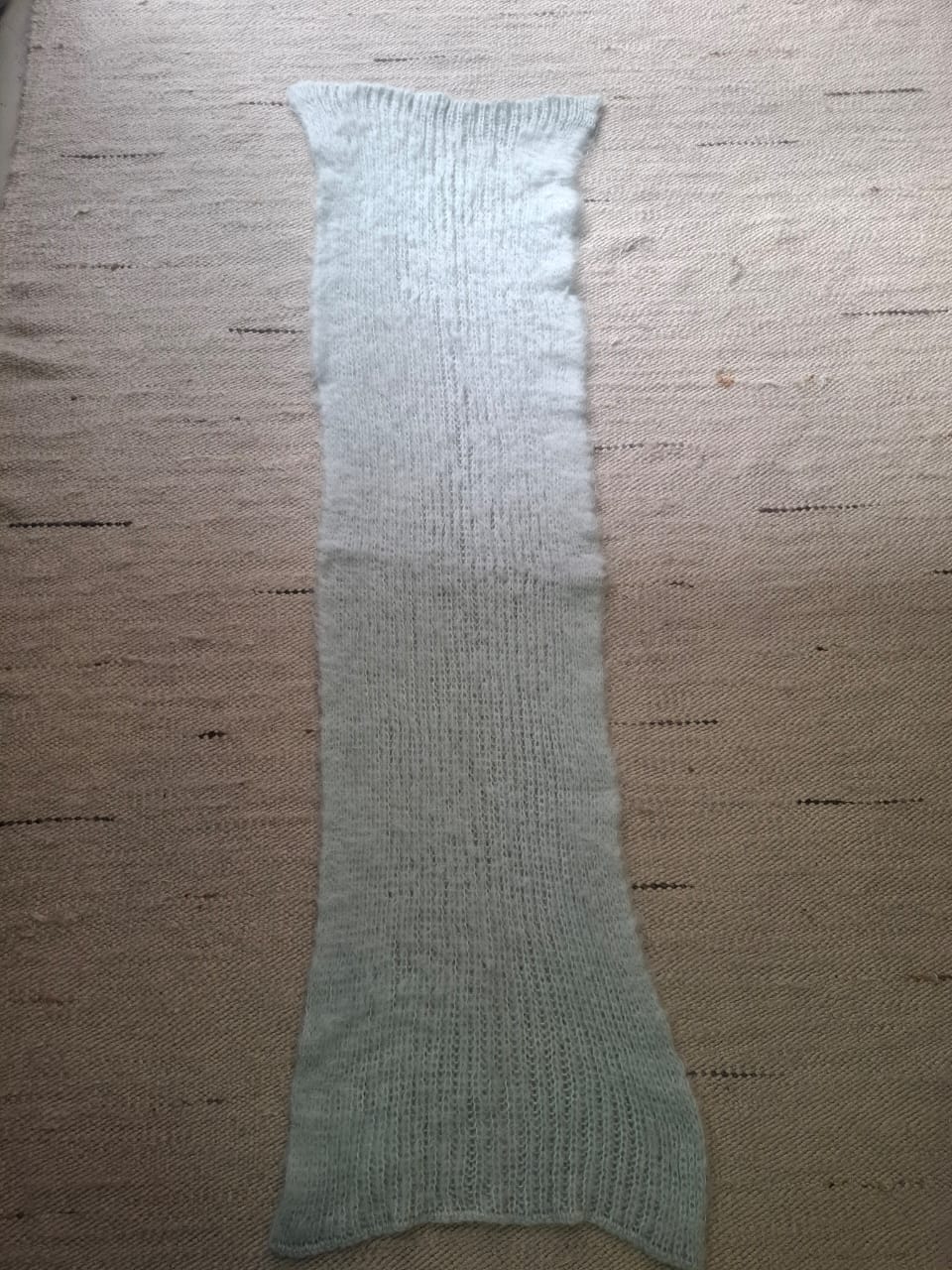 A long, narrow ribbed knit fabric piece in a Soft Mint  green color, laid flat on a textured beige woven rug.